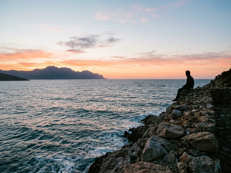 Person sitting on a rocky shoreline watching the sea and distant mountains at sunset