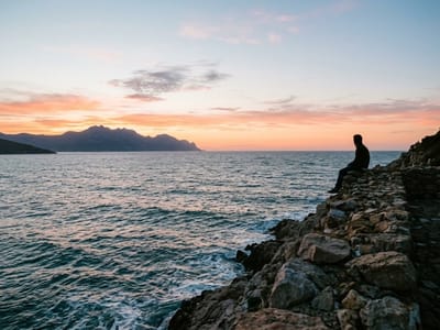Person sitting on a rocky shoreline watching the sea and distant mountains at sunset