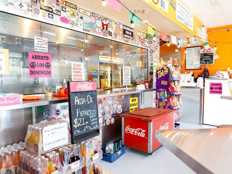 Counter-service interior of Las Tortas Perronas in Spring Branch, with a sticker-covered glass counter, chalkboard menu, and a red Coca-Cola cooler.