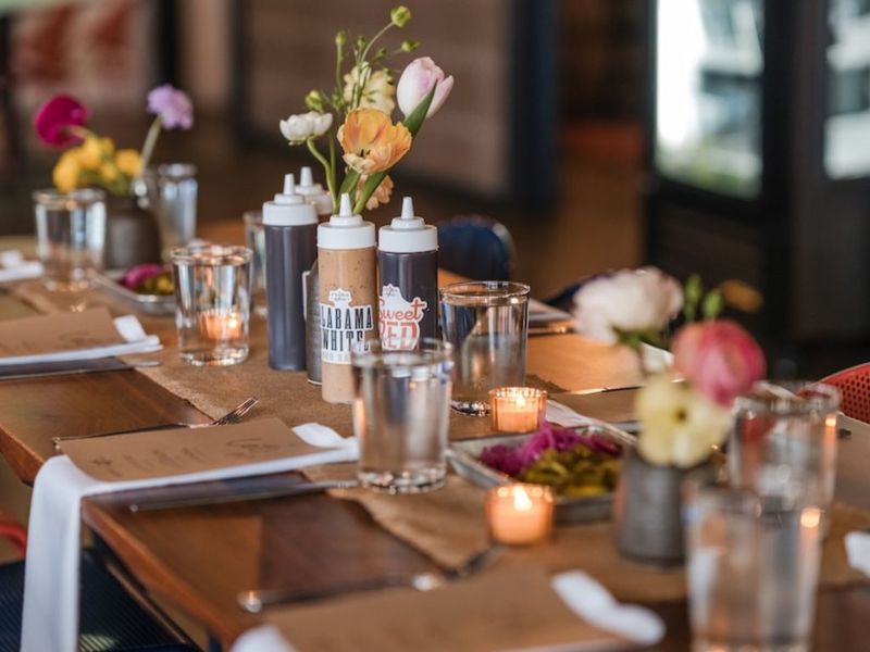 A set table at Feges BBQ with house sauce bottles, water glasses, votive candles, and a small vase of tulips.